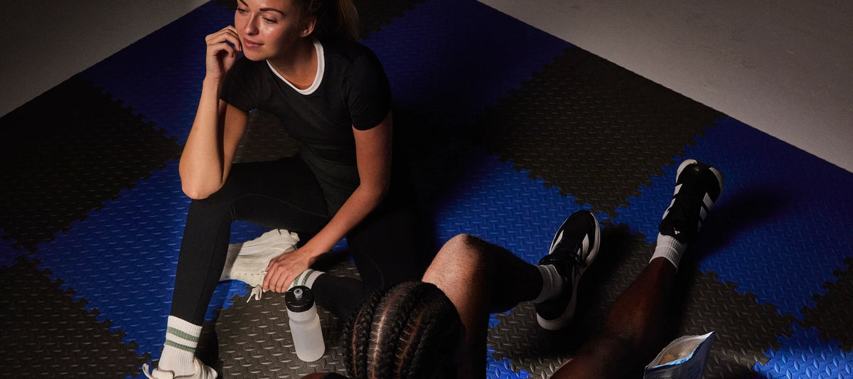 Two athletes resting on a gym floor with a bag of Subtle Snacks